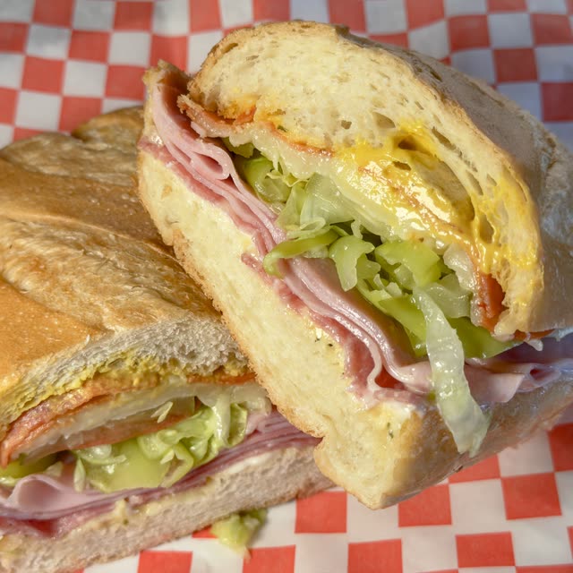 Italian Sub cut in half, layered with salami, pepperoni, ham, lettuce, tomato, and mustard on a soft roll