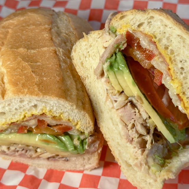 Skateboarder's Delight sandwich cut in half, with ham, turkey, bacon, provolone, avocado, tomato, and red onion on a soft roll